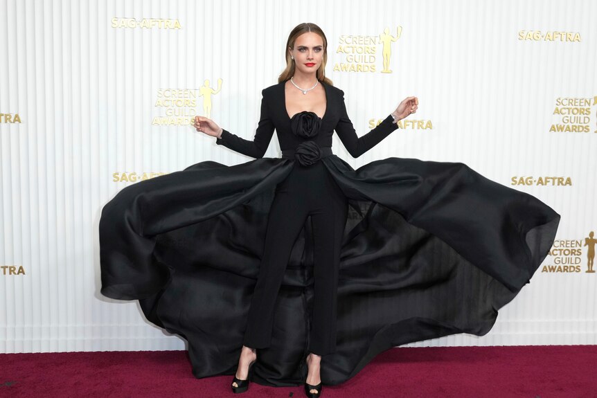 Cara Delevingne wearing a black pantsuit with a flowing black skirt blowing up around her