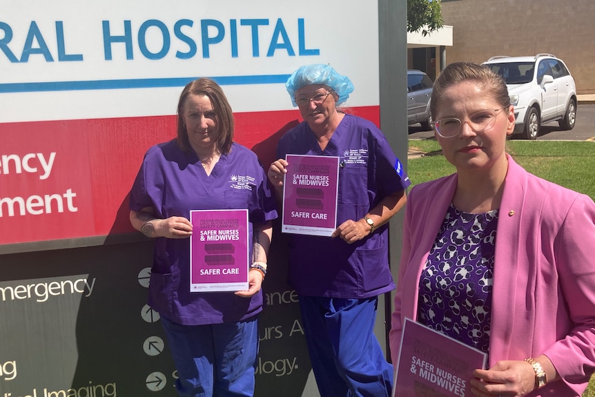 Three women standing in front of the hospital sign holding 'safer care' posters.