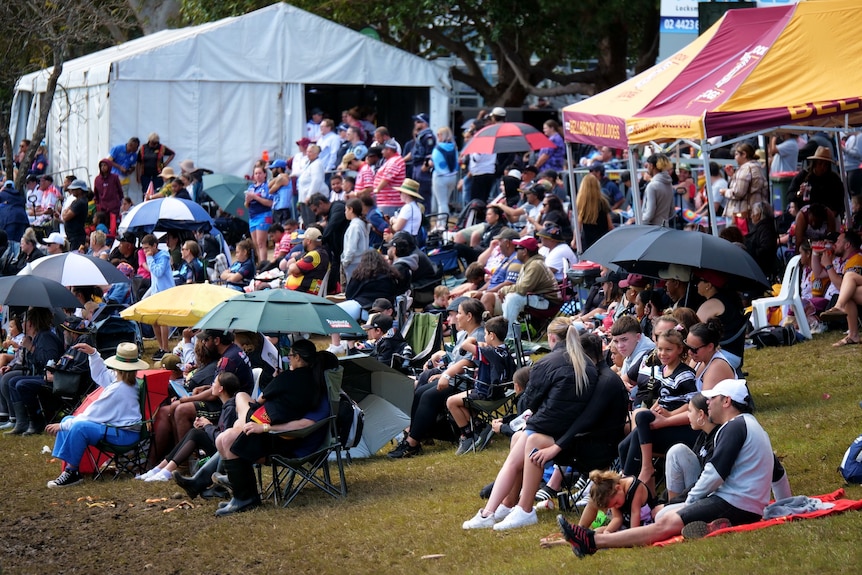 Crowd of Koori Knockout spectators sit in a grass hill with umbrellas and gazebos 