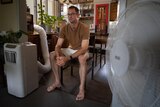 A man sits near an air conditioner unit and a pedestal fan.