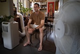 A man sits near an air conditioner unit and a pedestal fan.