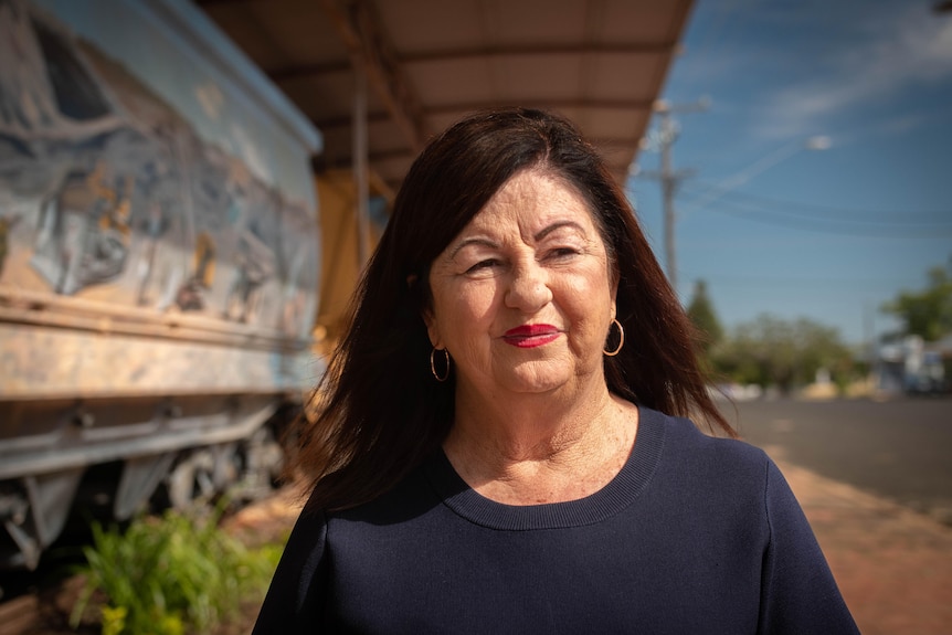 A woman with brown hair, bright red lipstick, stands on a sunny street staring into the distance.