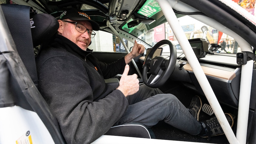 A man in the driver's seat of a car with a roll cage gives the thumbs up