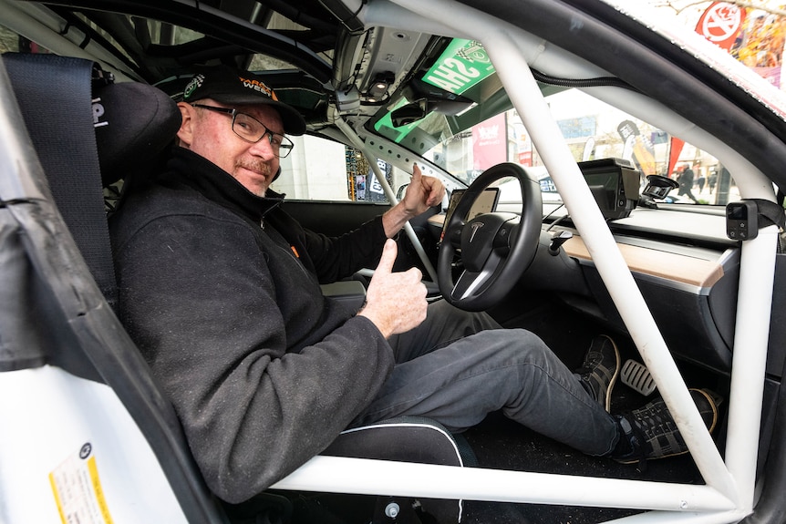 A man in the driver's seat of a car with a roll cage gives the thumbs up