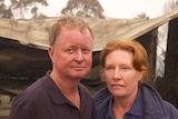 A man and a women put their arms around each other with a destroyed house behind them