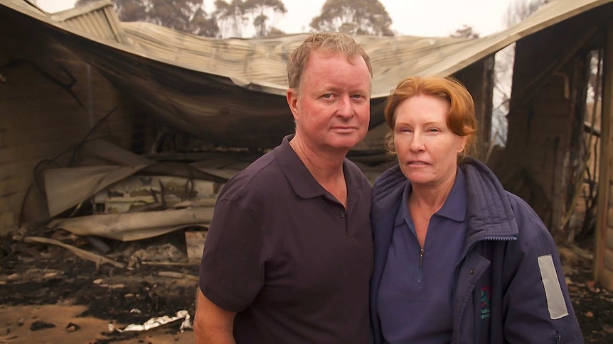 A man and a women put their arms around each other with a destroyed house behind them