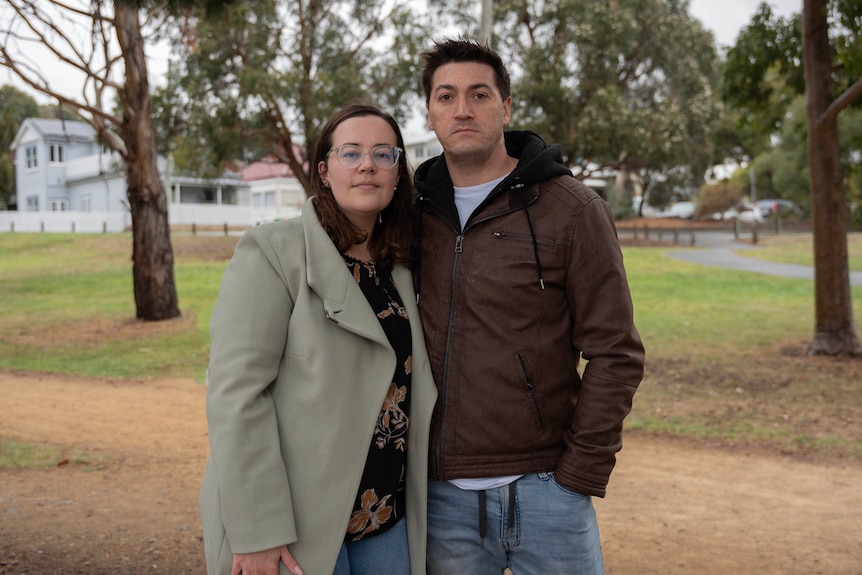 A man and woman stand in a park with a dirt track and house behind them.