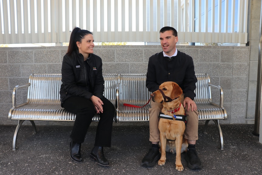 a woman and a man with an assistance dog sitting on a silver bench at a train station, smiling and talking to each other
