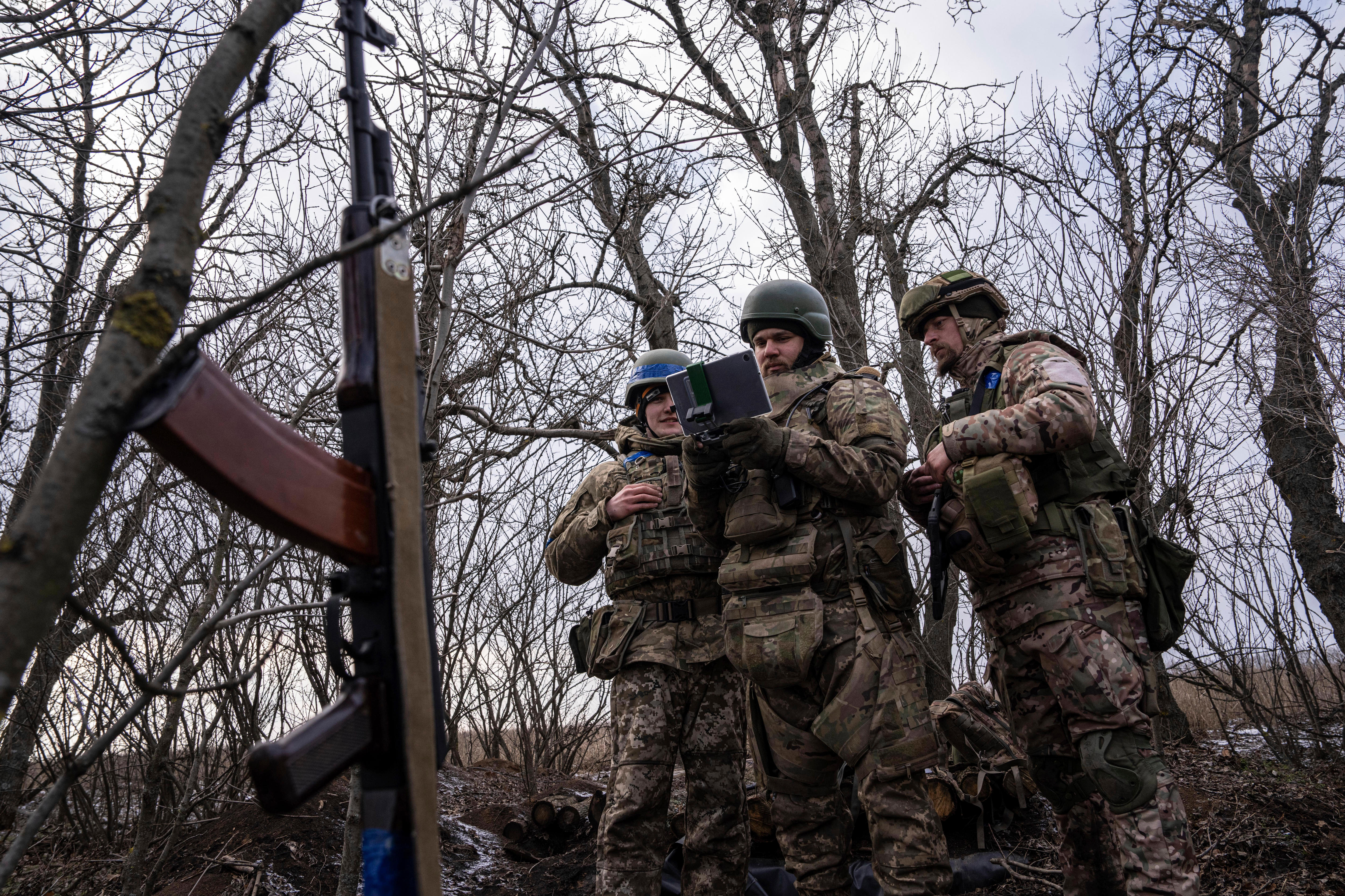 Soldiers gather in a stand of leafless trees to watch a drone operator in action with a smart device.