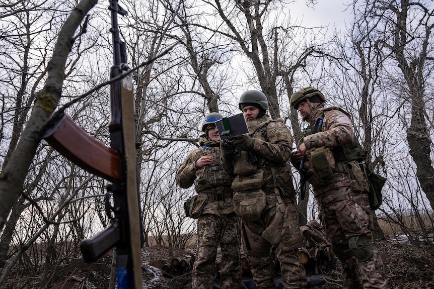 Soldiers gather in a stand of leafless trees to watch a drone operator in action with a smart device.