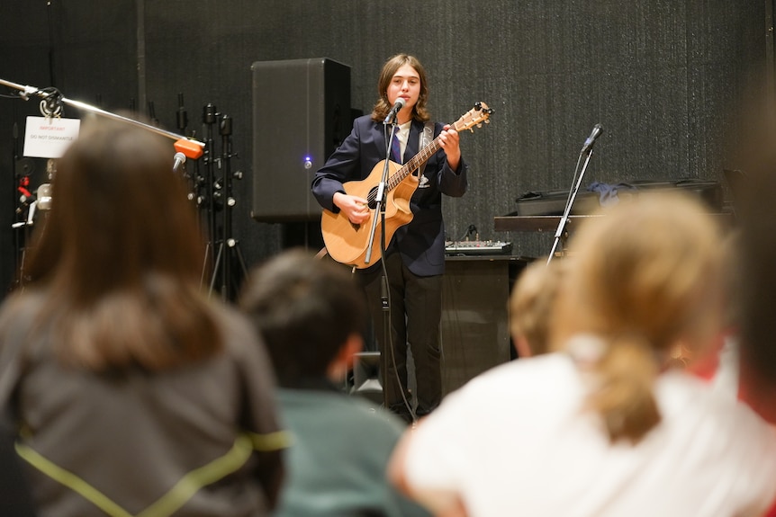 Rory stands in front of a microphone with a guitar. Children are seated on the floor in front of him.