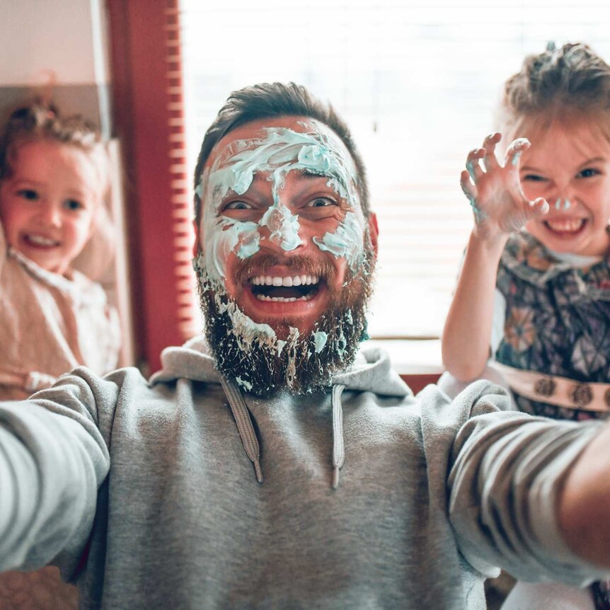 Dad in kitchen with kids, holding a camera with a grin and cake batter his on face, posing for a photo with two children