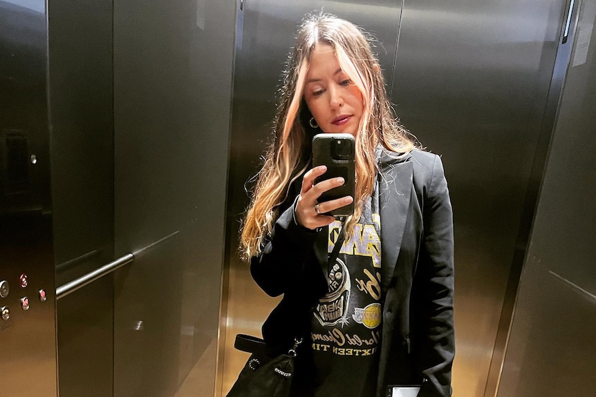 A woman in workwear takes a photo of herself in an elevator mirror.