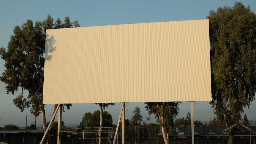 A drive in movie theatre screen, with trees visible behind it and a carpark in the foreground