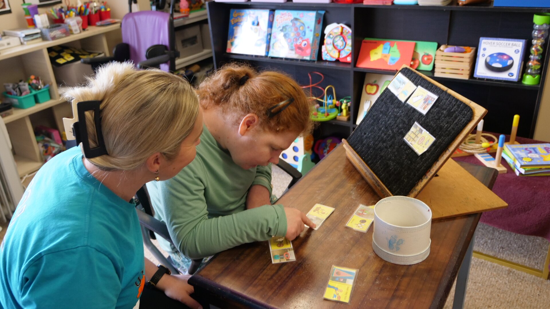 A young woman points to a card on her desk with a board they put cards onto and support worker sitting with her