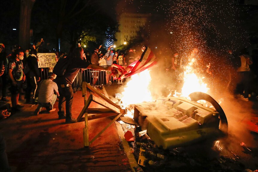 A man throws an American flag onto a fire that is surrounded by people filming with mobiles and hold protest signs.