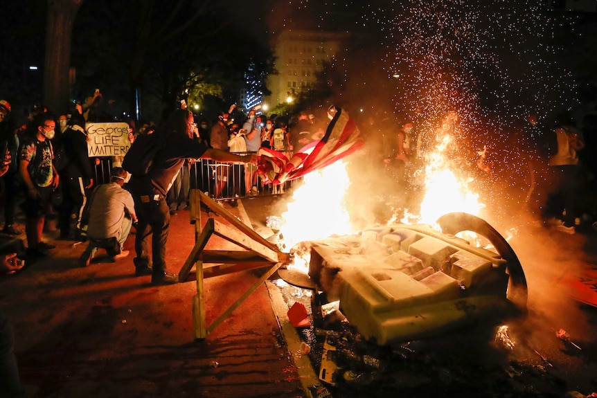 A man throws an American flag onto a fire that is surrounded by people filming with mobiles and hold protest signs.