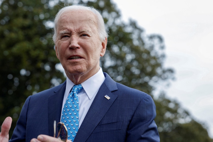 Image of Joe Biden talking, he is wearing a blue suit with a tie with tiny donkeys on it. Behind him are green leaves .
