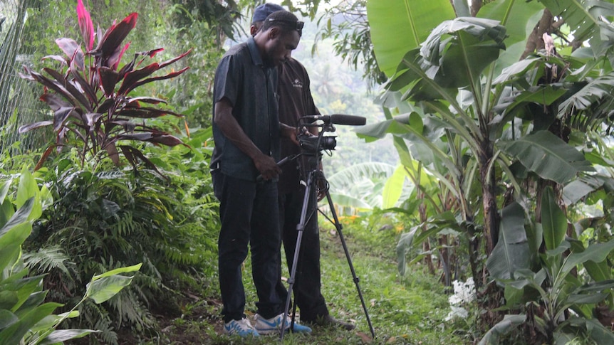 Two men with a camera on a tripod film the surrounding green bush