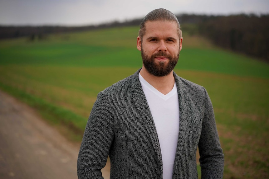 A bearded young man wearing a white shirt and grey jacket stands in the foreground with a blurred young wheat field in the back