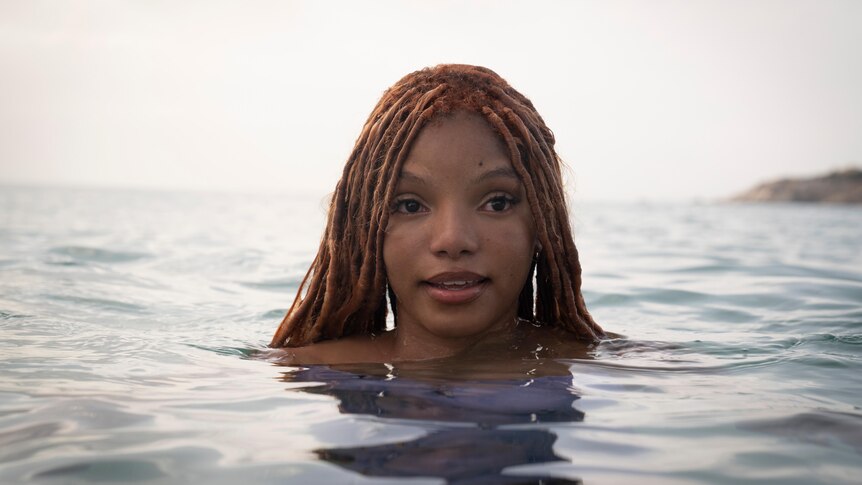A young black woman with red-toned dreadlocked hair swims in the sea, only her head visible above the water.,