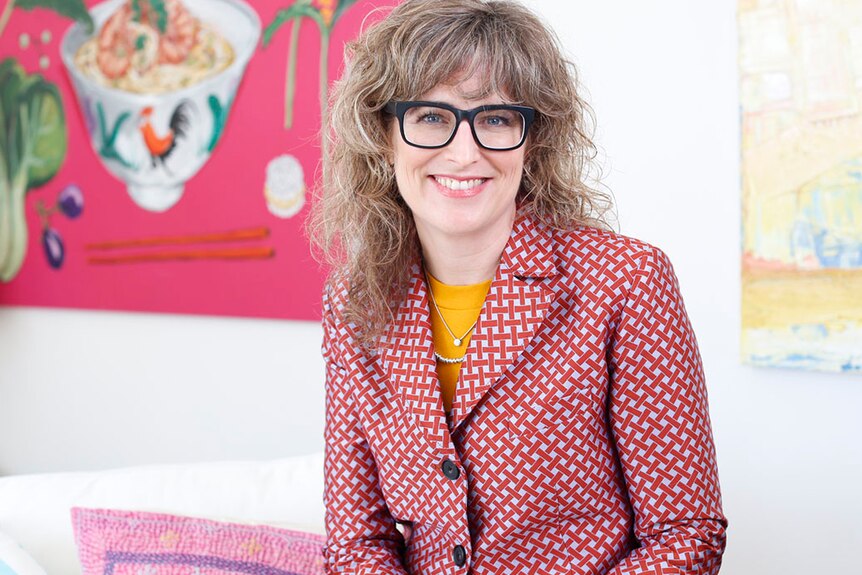 Laura Higgins wears a red, checkered jacket and sits in front of some bright modern paintings.