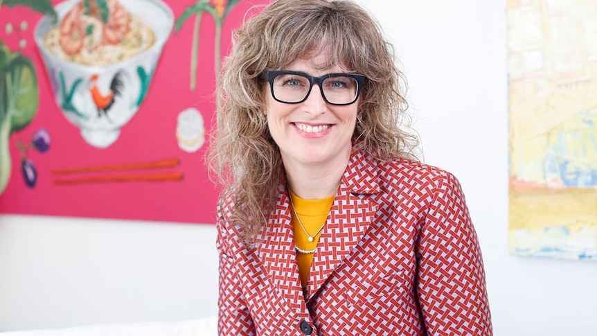 Laura Higgins wears a red, checkered jacket and sits in front of some bright modern paintings.