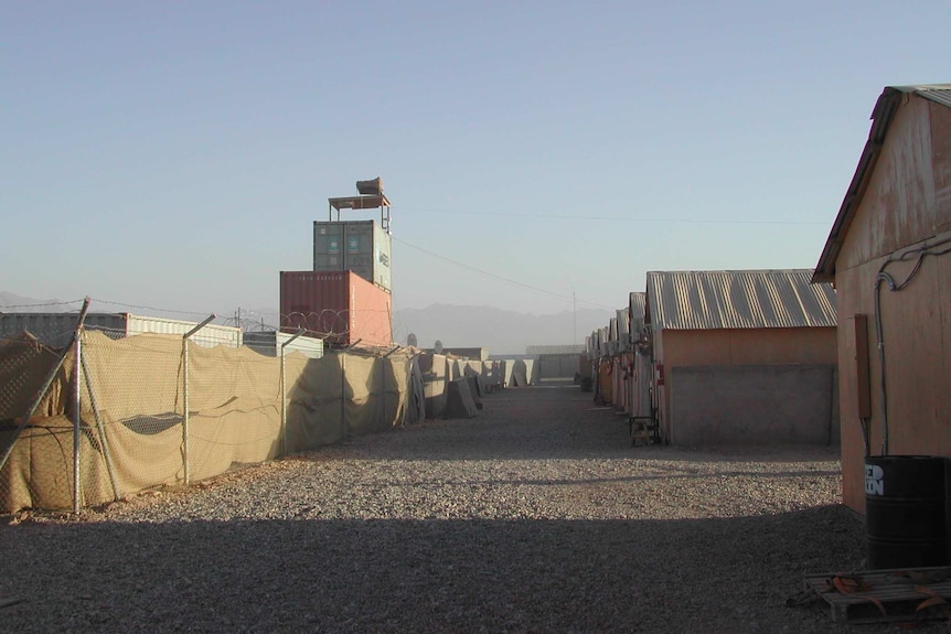 Multiple rows of plywood huts with iron roofs are separated by a dusty road. The air is dusty.