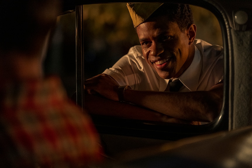 A night time a man wearing US Army garrison cap bends over and rests forearms on open car window and smiles at person inside.