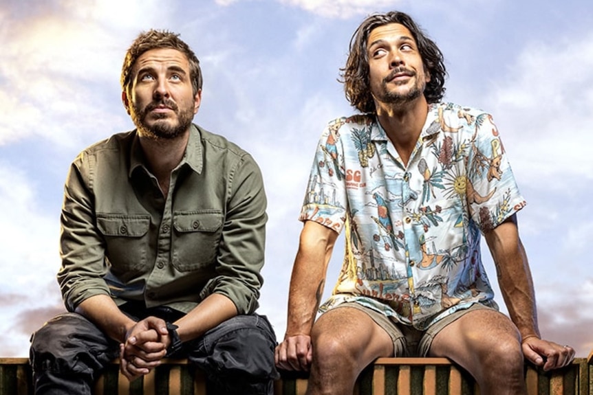 Two actors (Ryan Corr and Bob Morley) sitting on a ledge looking off into the distance, one in khaki green, the other in colours