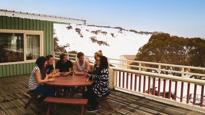 People sitting on a deck at a ski lodge with a snow-covered mountain in the background.
