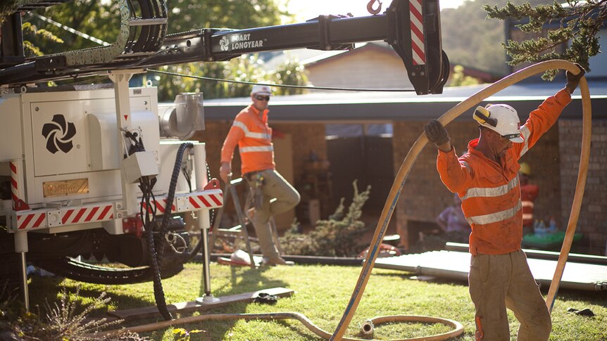 Two men in orange-coloured workwear at a bore drilling rig with house in background.