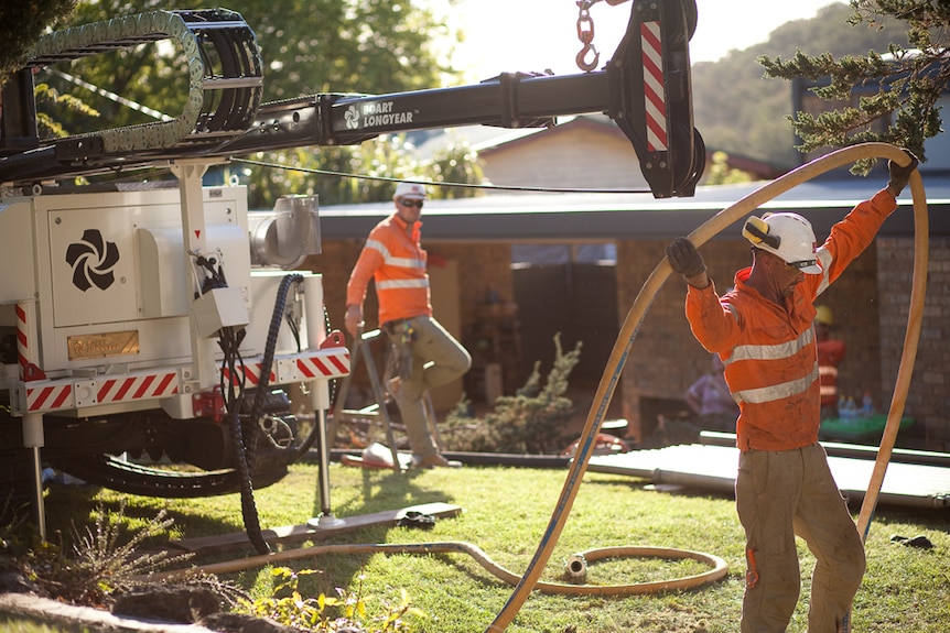 Two men in orange-coloured workwear at a bore drilling rig with house in background.