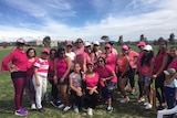 A dozen women in pink shirts pose around a cricket pitch.