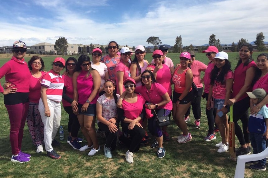 A dozen women in pink shirts pose around a cricket pitch.