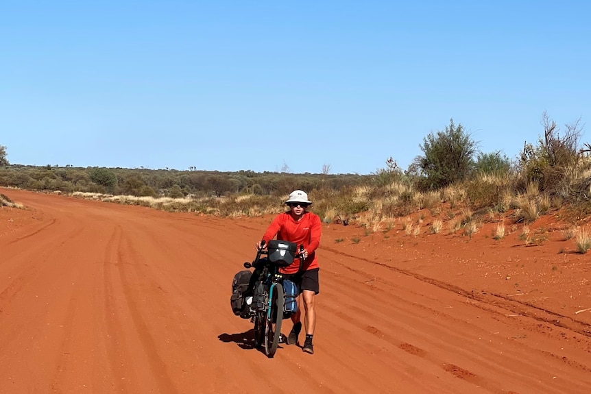 A young man walks with his bike along a dirt road in heavy sun protection gear. He looks exhausted with no clouds in sight.