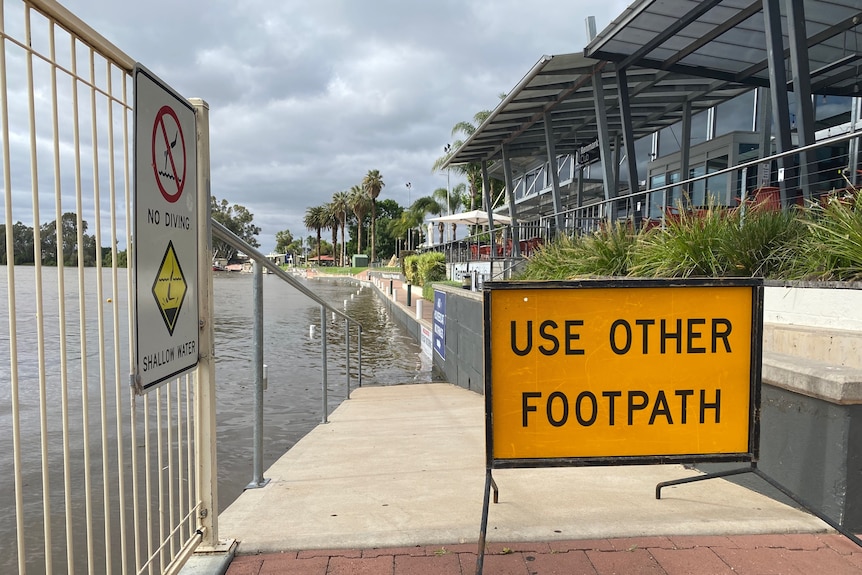 A sign which says use other footpath in front of a flooded footpath next to a river. 