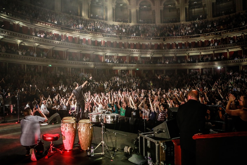 In a packed multi-tiered theatre a crowd of people raising their hands faces a stage on which Felix Reibl raises his mic up.