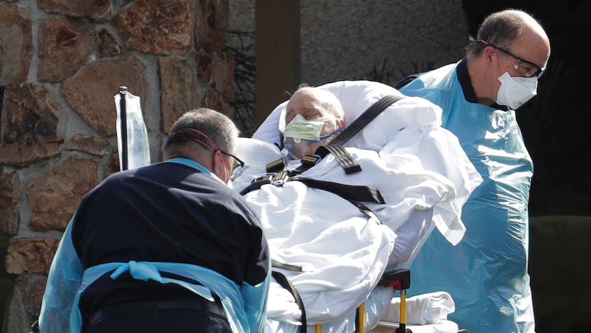 An old man with eyes closed and with face mask is wheeled on bed by two men wearing scrubs and masks