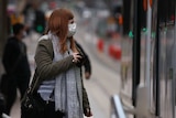 A woman with long copper-coloured hair stands at a tram stop while wearing a mask and scarf in Melbourne.