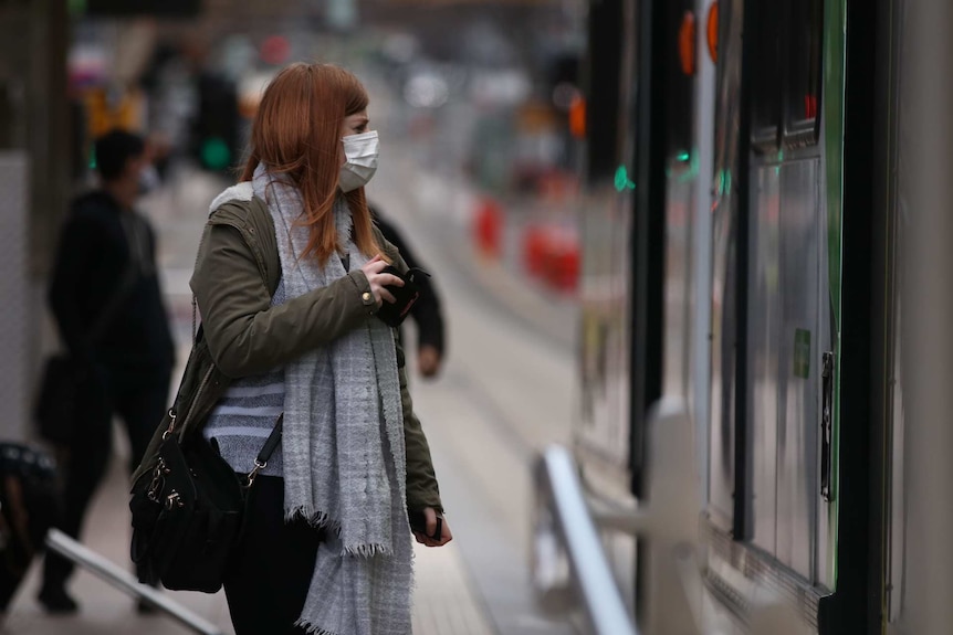 A woman with long copper-coloured hair stands at a tram stop while wearing a mask and scarf in Melbourne.