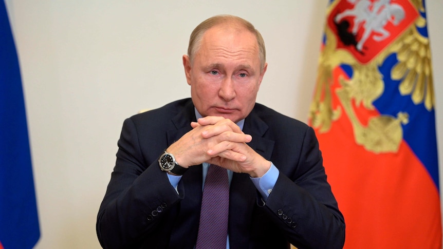 Man wearing a suit sitting at a desk with the Russian flag behind him. 