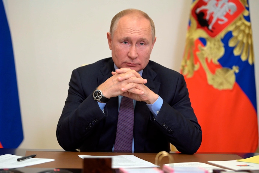 Man wearing a suit sitting at a desk with the Russian flag behind him. 