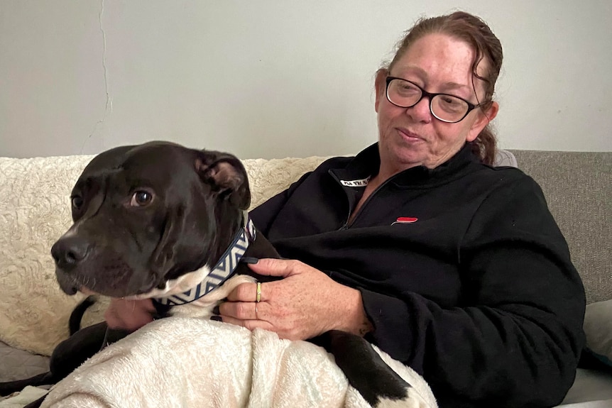A Caucasian lady with brown hair, black glasses and black jumper cuddles her black and white dog on a grey sofa