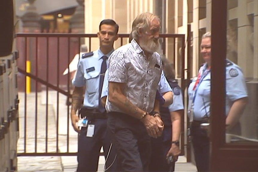 A grainy image of an older man with a white beard walking outside of a court, his hands are cuffed.