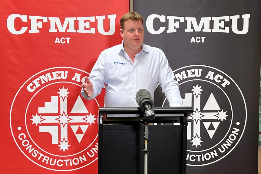 A man stands at a lectern with signs for CFMEU behind him.