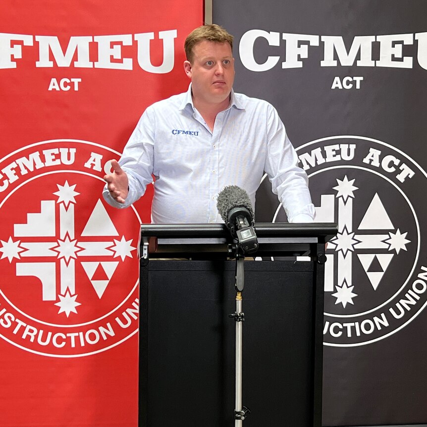 A man stands at a lectern with signs for CFMEU behind him.
