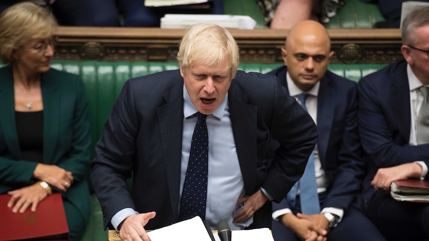 Boris Johnson stands at the lectern to speak in the House of Commons. He is mid-speech and wears a serious expression.