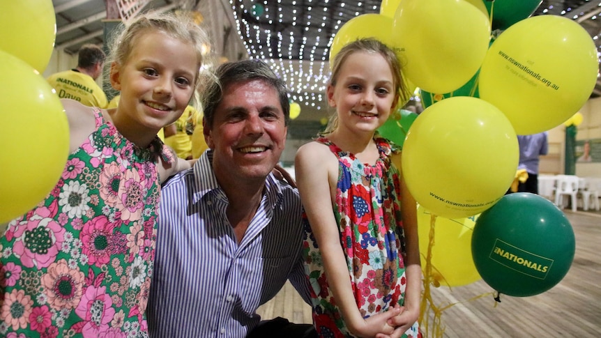 A man squats down beside his two young daughters surrounded by yellow balloons.