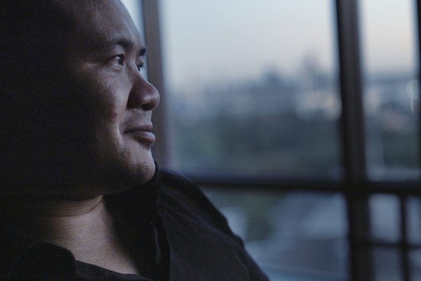 A man in a black shirt sits in the dark in front of a window.